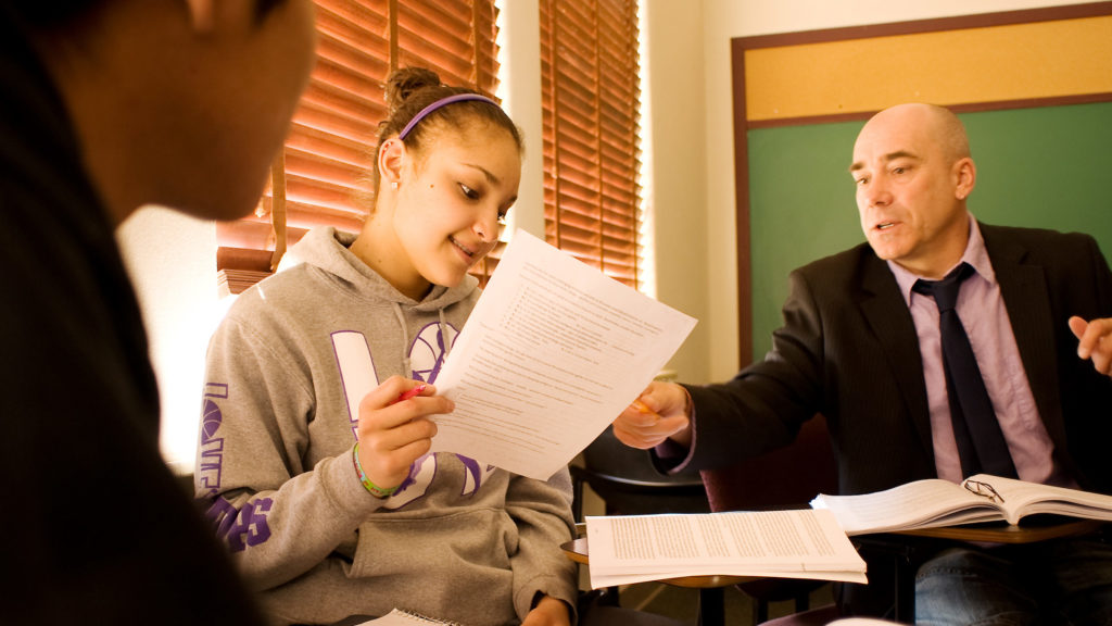 bald teacher helping female student with essay while male student looks over essay.